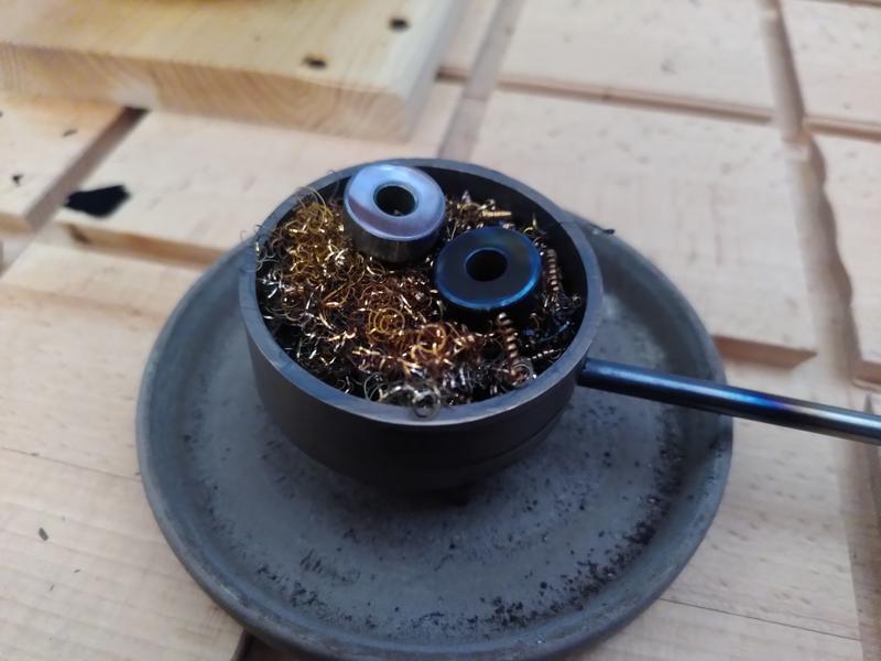 A round brass blueing tray filled with bronze chips. The blueing tray
has a steel handle going off to the right. The blueing tray is sat in a
ceramic dish. On top of the chips two steel parts. One is a shiny dark
blue. One a shiny steel colour.